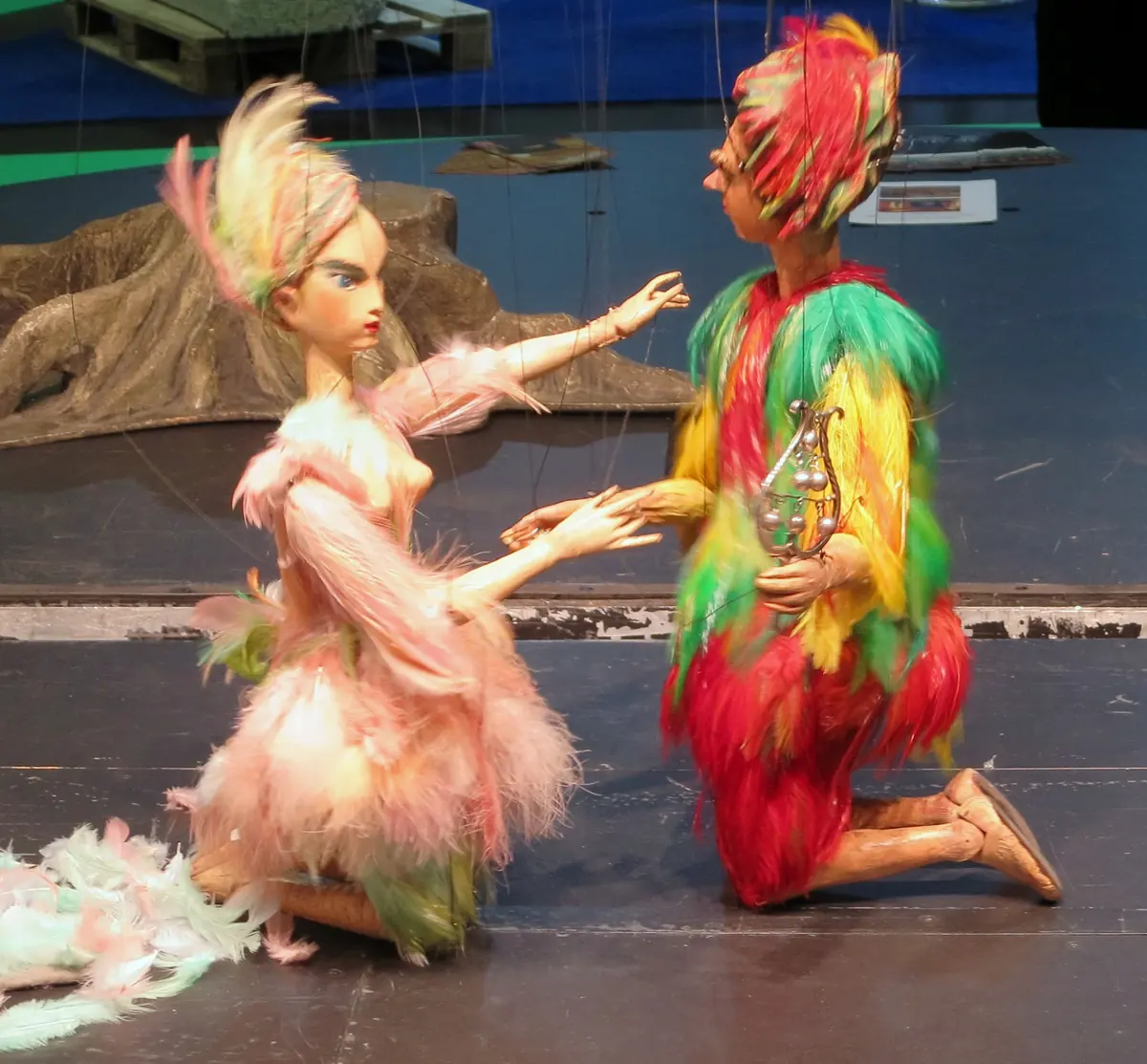Two marionettes in feathered costumes, one pink and one red, yellow and green, kneel facing each other on a stage, with fine strings visible above them