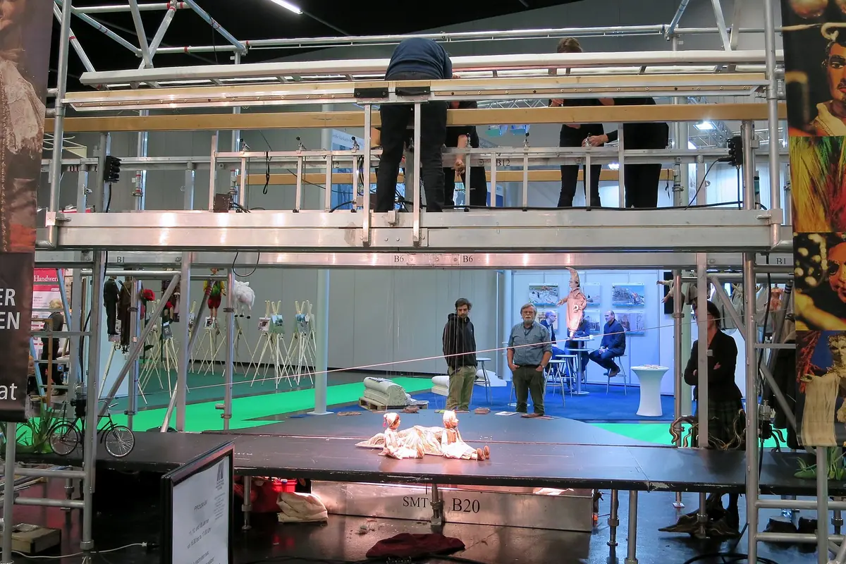 A metal scaffold platform above a small stage, with people working on the upper level and a marionette lying on the stage below