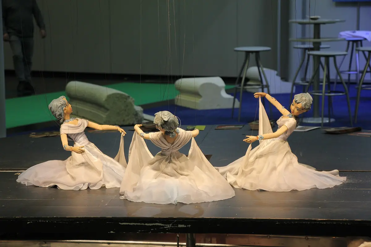 Three marionettes in long white gowns kneel and lean on a dark stage with strings above them