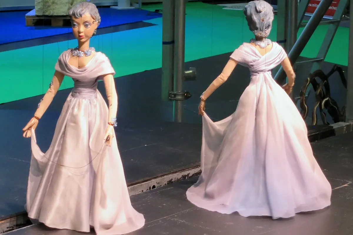 Two marionettes in long pale gowns hold out their skirts on a stage, one seen from the front and one from behind