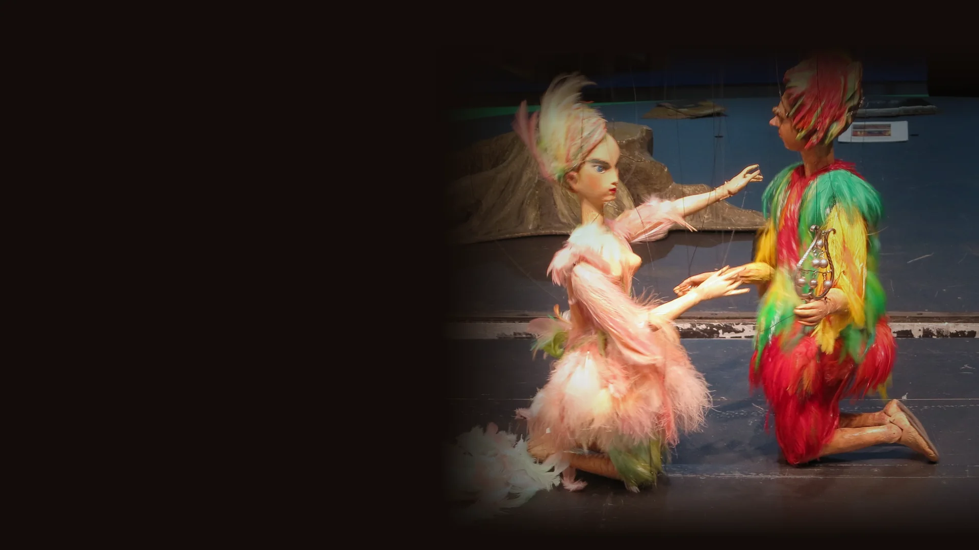 Two marionettes in feathered costumes, one pink and one red, yellow and green, kneel facing each other on a spotlit stage, with fine strings visible above them